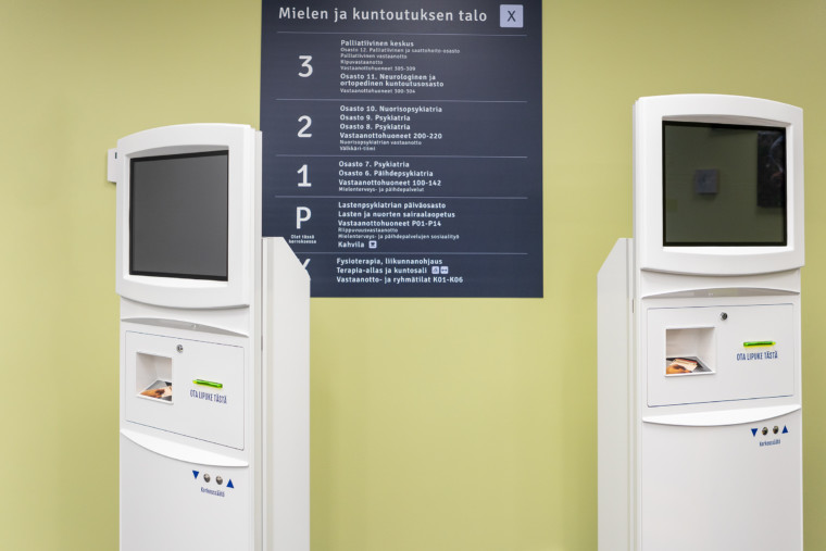 Kaksi valkoista ilmoittautumisautomaattia, seinällä opastetaulu.