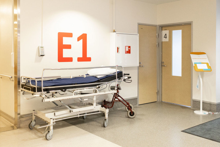 Terveyskeskuksen käytävä, jonka seinustalla tyhjä potilasvuode ja rollaattori. Seinällä suurin kirjaimin E1