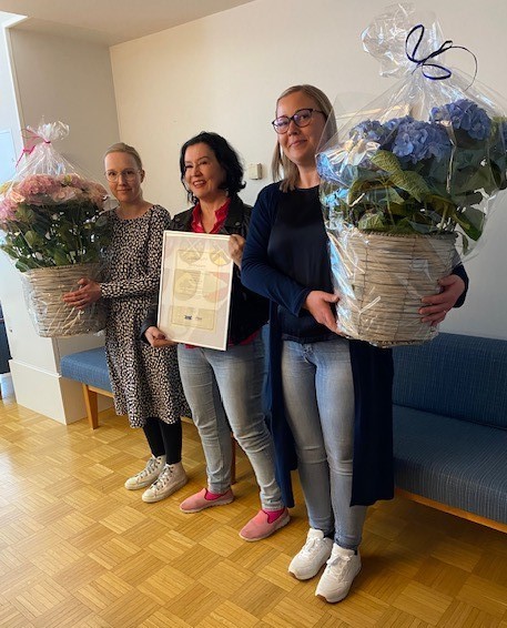 Maarit Rantakurtakko pitää sertifikaattia hymyillen. Hänen molemmilla puolilla seisovat Piia Suihkonen ja Heidi Laurila isot kukkapuskat käsissään.