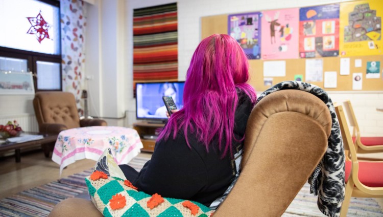 Pinkkitukkainen nainen istuu nojatuolissa selin kameraan katsomassa televisiota