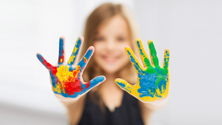 kuva tytön kämmenistä, joissa on eri värisiä sormivärimaaleja, taustalla sumeana tytön hymyilevät kasvot