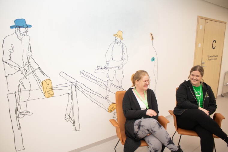 Eloisan ammattilaiset Satu Vuorio ja Lotta Nokelainen istuvat tuoleilla, katsovat toisiaan ja nauravat. Kuva on otettu Savonlinnan sote-keskuksen käytävällä. Taustalla näkyy seinälle tehty taideteos kahdesta piirretystä miehestä, jotka sahaavat puita.