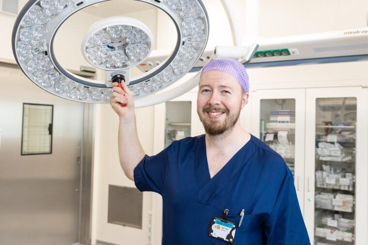 siniasuinen mieskirurgi leikkausalissa katsoo kameraan ja säätää kädellään leikkausvalaisinta