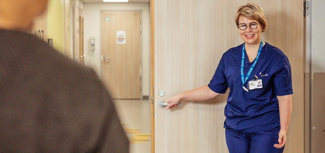 siniasuinen sairaanhoitaja terveyskeskuksen vastaanottohuoneen ovella ja käytävää pitkin selin kameraan päin kävelevä henkilö
