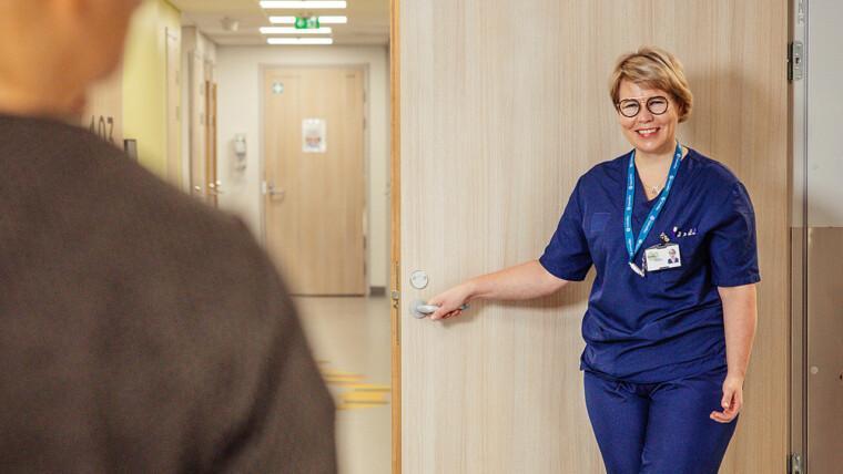 siniasuinen sairaanhoitaja terveyskeskuksen vastaanottohuoneen ovella ja käytävää pitkin selin kameraan päin kävelevä henkilö
