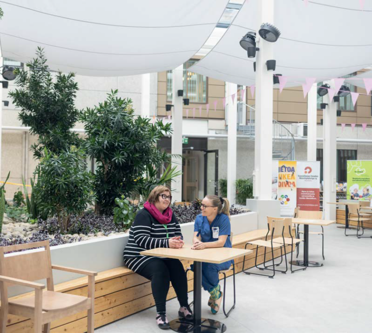 Kuvassa Tiina Aalto ja Tiia Kokkonen istumassa Savonlinnan keskussairaalan kansalaistorilla pöydän ääressä. Tiina ja Tiia keskustelevat ja hymyilevät toisilleen.