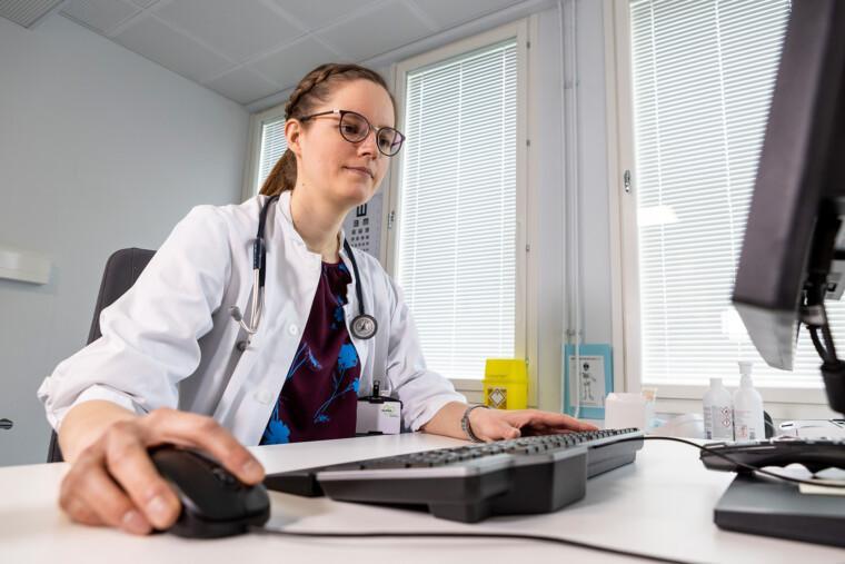 naislääkäri valkoisessa takissa istuu pöydän ääressä käyttäen tietokoneen näppäimistöä ja hiirtä