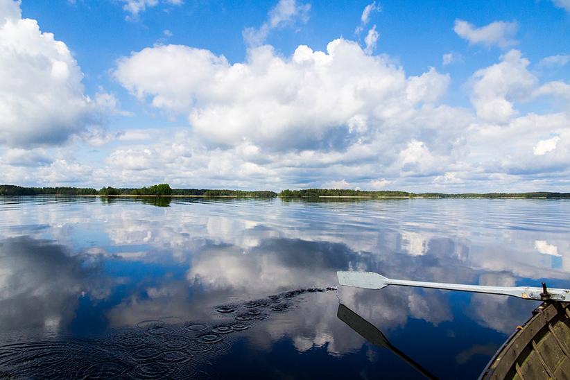 Kuvituskuva: Kesäinen järvimaisena, jossa taivas heijastuu järvenpinnasta. Veden yllä on soutuveneen airo ja kulmassa näkyy hieman veneen laitaa.