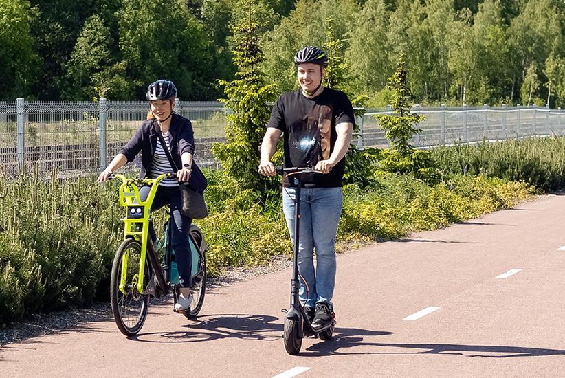 Pyöräilijä ja sähköpotkulautailija ajavat pyörätiellä. Molemmilla on kypärät päässä.