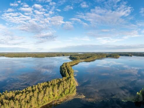 Ilmakuva saimaalta. Vettä, sininen taivas jossa hieman pilviä. Keskellä kiemurtelee taigametsäinen kaistale.