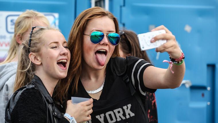 Kaksi nuorta tyttöä ottavat selfietä festareilla ja ilmeilevät kameralle.