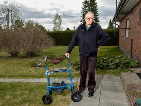 Heikki seisoo asuntonsa pihalla ja hän pitää kevyesti kiinni vieressään olevasta rollaattorista.
