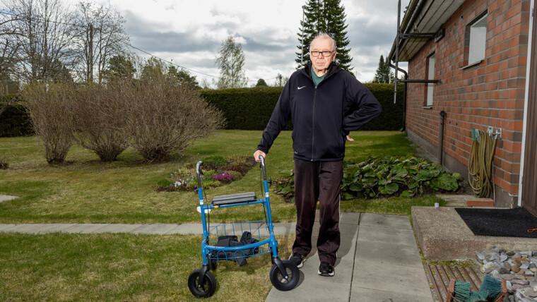Heikki seisoo asuntonsa pihalla ja hän pitää kevyesti kiinni vieressään olevasta rollaattorista.