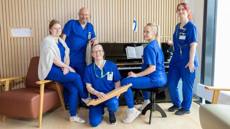 Neljä naista ja yksi mies kuvassa. Osa istuu ja osa seisoo. Kaikki hymyilevät kameralle. Yksi istuu pianon edessä pianojakkaralla ja yksi pitää käsissään kanteletta.