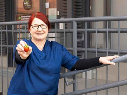 Maija seisoo työvaatteissaan sote-aseman edessä ja nojaa kaiteeseen. Maija hymyilee ja pitää kädessään mansikkaa, jota ojentaa kameralle.
