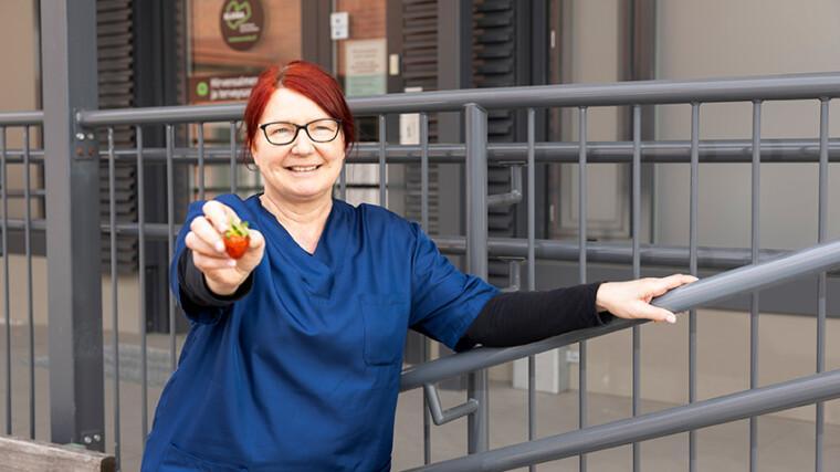 Maija seisoo työvaatteissaan sote-aseman edessä ja nojaa kaiteeseen. Maija hymyilee ja pitää kädessään mansikkaa, jota ojentaa kameralle.