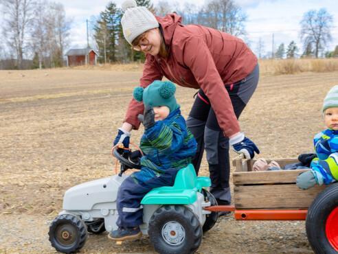 Lapsi istuu lasten polkutraktorissa ja vetää pientä kärryä. Kärryssä istuu toinen lapsi. Nainen kumartuu traktorissa olevan lapsen puoleen ja hymyilee. On alkukevät, kaikilla on vielä talvivaatteet päällään.