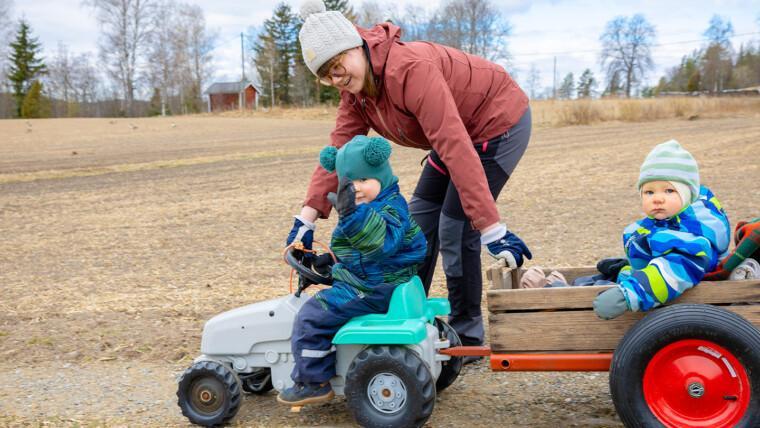 Lapsi istuu lasten polkutraktorissa ja vetää pientä kärryä. Kärryssä istuu toinen lapsi. Nainen kumartuu traktorissa olevan lapsen puoleen ja hymyilee. On alkukevät, kaikilla on vielä talvivaatteet päällään.