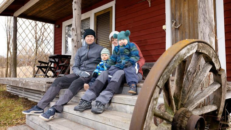 Vanhemmat istuvat vanhan, punaiseksi maalatun talon portailla kaksi lasta sylissään. Etualalla näkyy vanhaa kärryn pyörää.