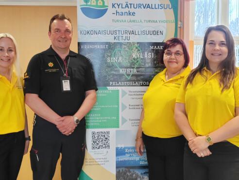 Kolme naista ja yksi mies seisovat Kylä turvallisuus-hankkeen banderollin edessä ja hymyilevät kameralle. Miehellä on päällään pelastuslaitoksen paita. Naisilla on päällä keltaiset järvi-suomen kylät ry:n paidat.