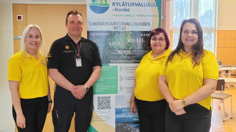 Kolme naista ja yksi mies seisovat Kylä turvallisuus-hankkeen banderollin edessä ja hymyilevät kameralle. Miehellä on päällään pelastuslaitoksen paita.
