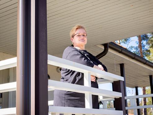 Laura nojaa kädet eteen ristittynä terassin kaiteeseen ja hymyilee kameralle. Aurinko paistaa. Lauralla on vaaleat lyhyet hiukset ja silmälasit.
