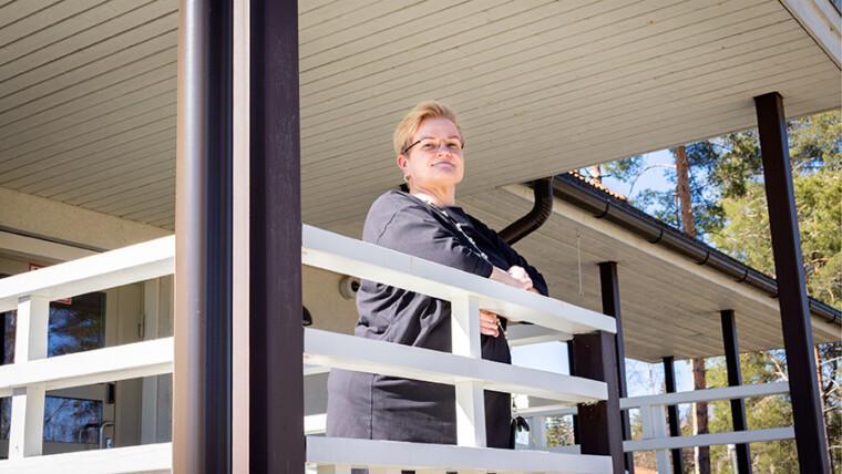 Laura nojaa kädet eteen ristittynä terassin kaiteeseen ja hymyilee kameralle. Aurinko paistaa. Lauralla on vaaleat lyhyet hiukset ja silmälasit.