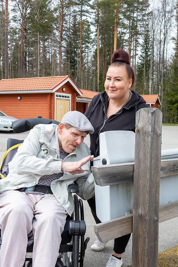 Mies istuu pyörätuolissa ja kurottuu katsomaan avoinna olevaan postilaatikkoon. Hänen takanaan seisoo nainen, joka pitää postilaatikon kantta auki ja hymyilee kameralle.
