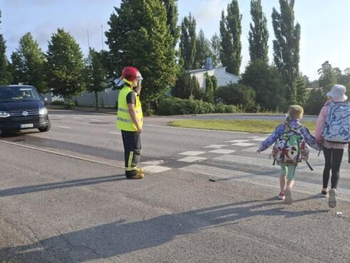 Kuvituskuvassa koululaiset reput selässä ylittävät suojatietä. Sopimuspalokuntalainen seisoo suojatien vierellä huomioliivit päällä näyttämässä pysäytysmerkkiä autoilijalle, joka on pysähtynyt suojatien taakse.