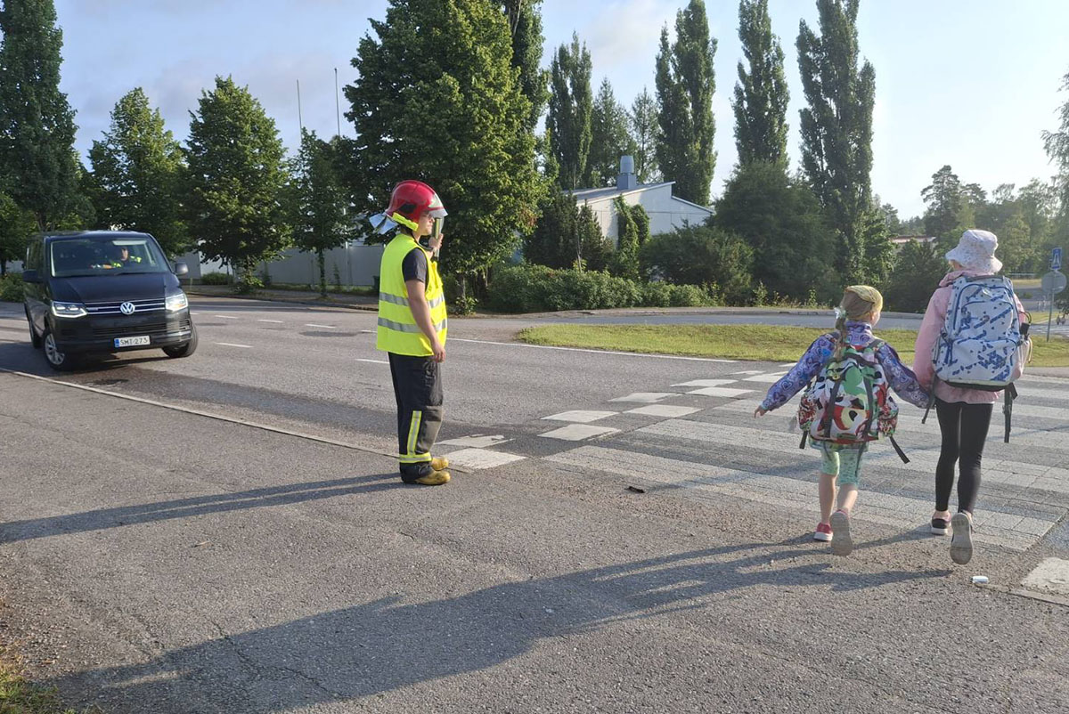 Kuvituskuvassa koululaiset reput selässä ylittävät suojatietä. Sopimuspalokuntalainen seisoo suojatien vierellä huomioliivit päällä näyttämässä pysäytysmerkkiä autoilijalle, joka on pysähtynyt suojatien taakse.
