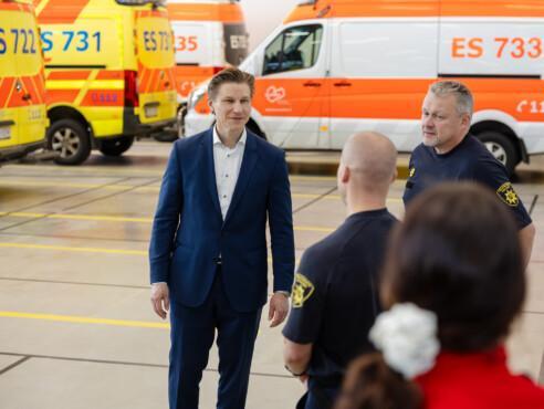 Ministeri paloaseman kalostohallissa henkilökunnan kanssa, taustalla ambulansseja.