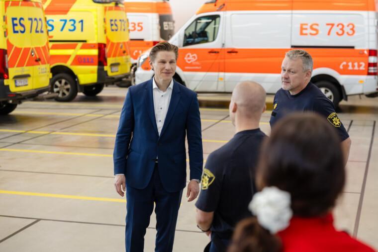 Ministeri paloaseman kalostohallissa henkilökunnan kanssa, taustalla ambulansseja.
