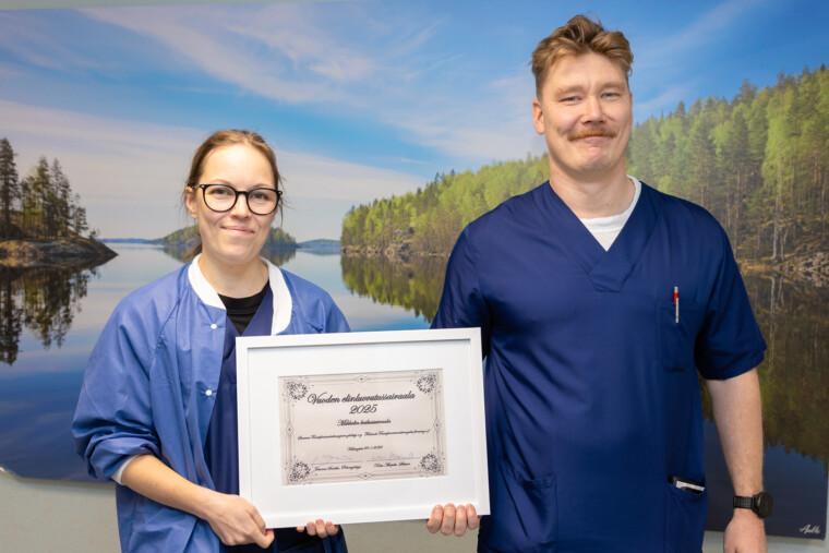 Siniasuiset sairaanhoitaja ja lääkäri pitävät käsissään kehystettyä kunniakirjaa