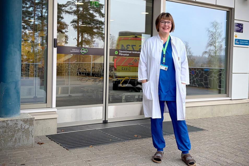 Valkoiseen lääkärintakkiin pukeutunut nainen seisten ja hymyillen Savonlinnan sairaalan pääsisäänkäynnin edustalla, taustalla olevista lasi-ikkunoista heijastuu pihalla oleva ambulanssi.